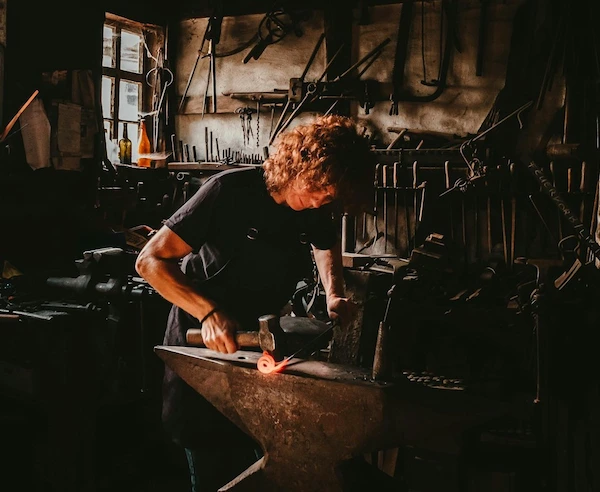 man står i en smedja och smider en metallbit med hammare